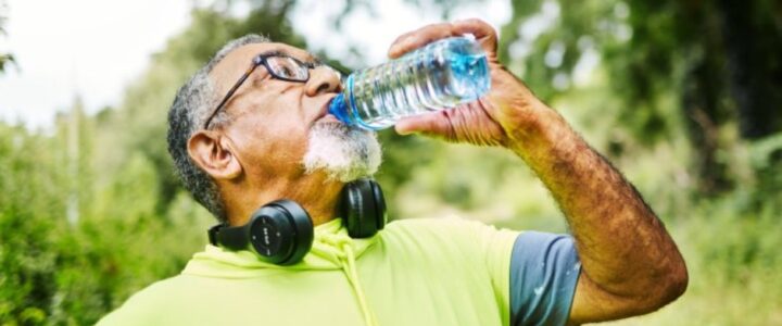A senior man wearing glasses, headphones around his neck, and fitness gear drinks from a water bottle on a wooded trail.