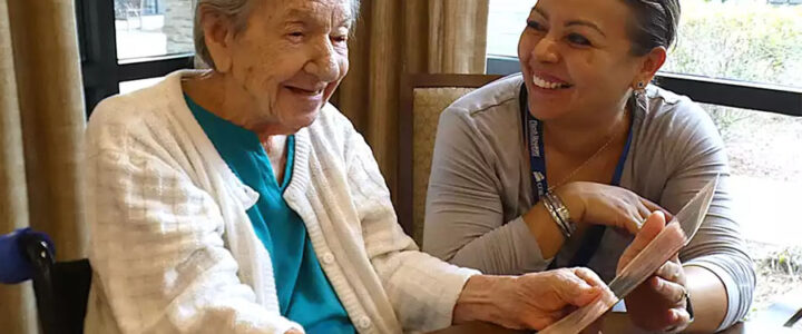 Elderly woman smiling and looking at a paper in her hands with a caretaker by her side.