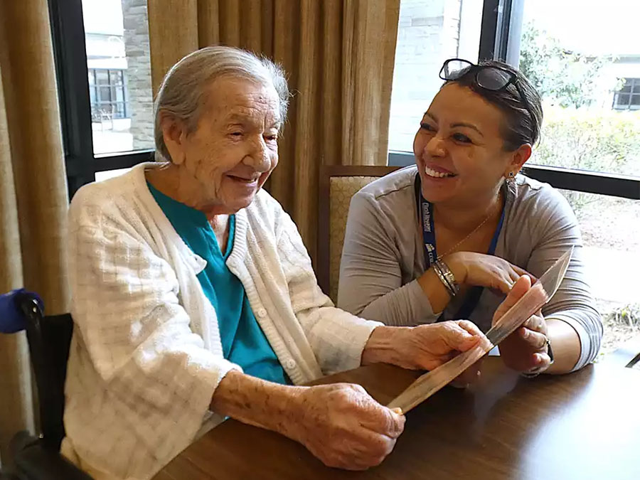 Elderly woman smiling and looking at a paper in her hands with a caretaker by her side.