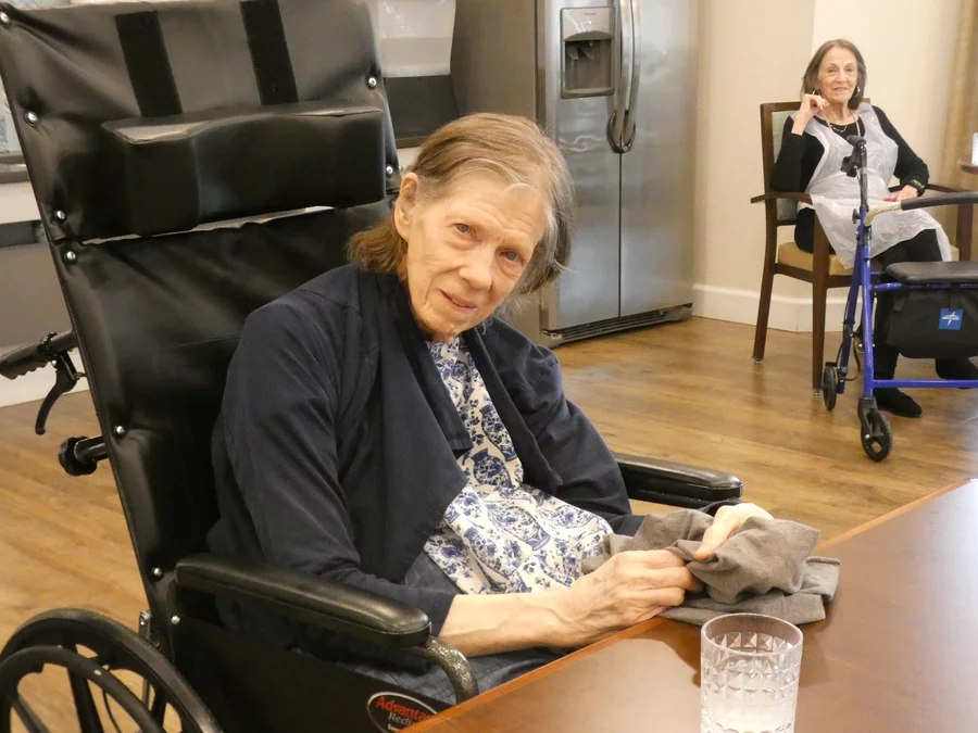 An elderly woman with a black cardigan over a blue and white patterned shirt.
