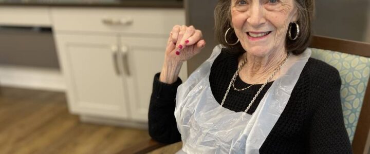 An older woman smiling in a kitchen with a plastic apron over her clothes.