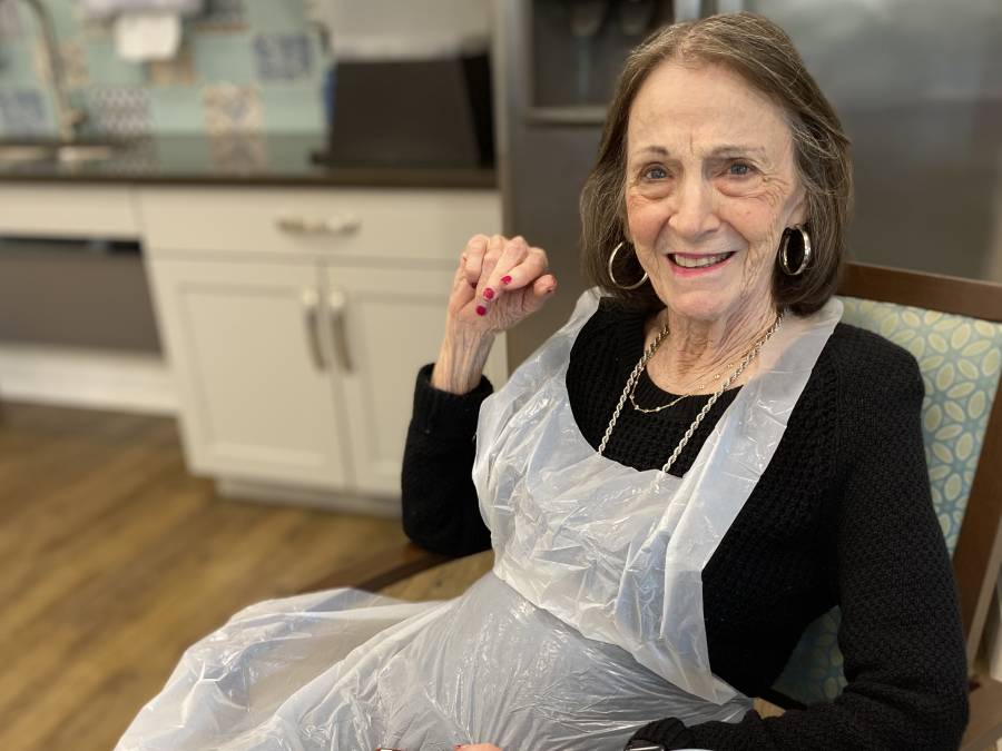 An older woman smiling in a kitchen with a plastic apron over her clothes.