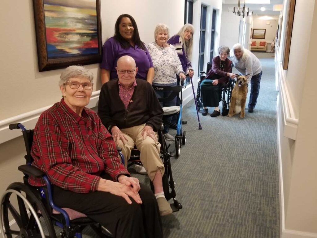 Seniors using mobility aids to stand and sit together in an assisted living hallway.