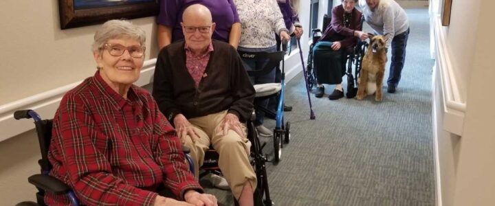 Seniors using mobility aids to stand and sit together in an assisted living hallway.