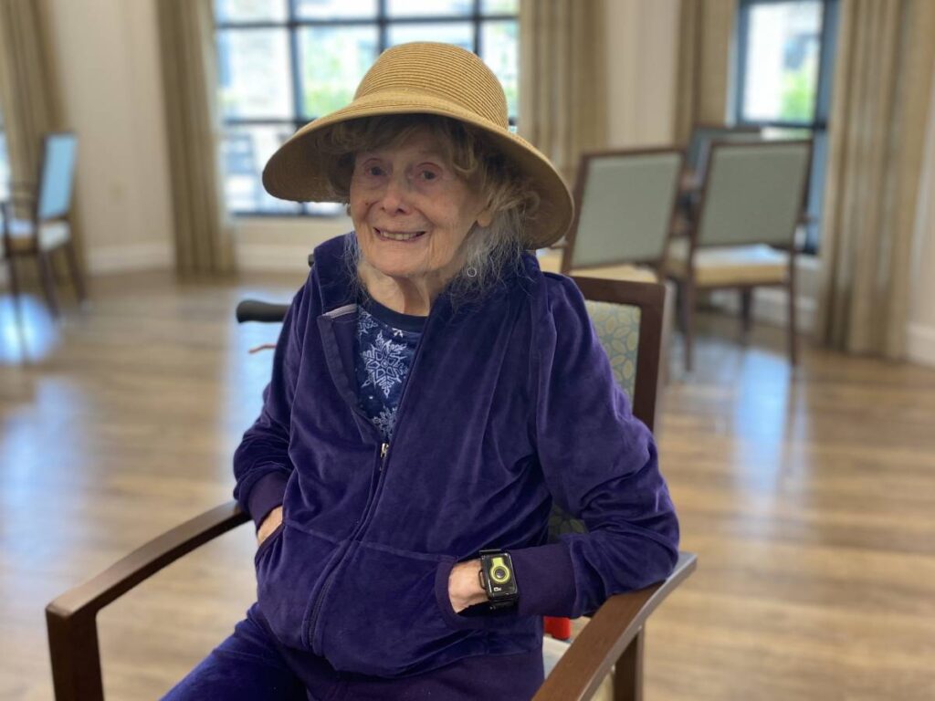 A senior woman in a memory care community wearing a navy velour tracksuit and straw hat.