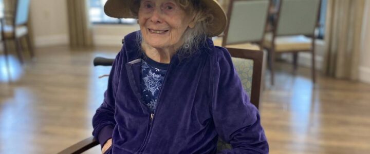 A senior woman in a memory care community wearing a navy velour tracksuit and straw hat.
