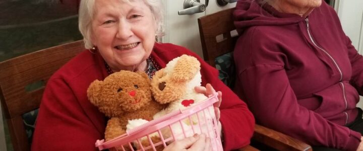Two senior women in red sweaters, one holding a basket of Valentine’s teddy bears.